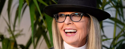 Diane Keaton in a tall black hat, dress shirt and dark blazer smiling in front of plants