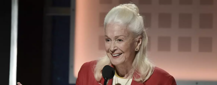 Diane Ladd in a red blazer and white scarf holding her hands out as she speaks into a microphone on a stage. Diane Ladd in a red blazer and white scarf holding her hands out as she speaks into a microphone on a stage.