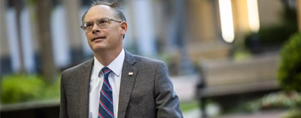 A man wearing glasses, a gray jacket and striped blue and red tie looks up in an outdoor setting