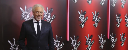 Tom Jones in a suit standing in front of a backdrop with The Voice UK logo