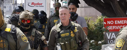 A man in an olive green uniform and vest walks with other people in similar uniforms near buildings