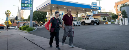 Two people walk on a sidewalk next to a gas station.