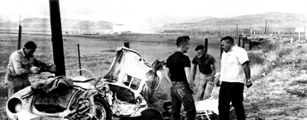 A black and white photo showing several men examining the remains of a wrecked sports car