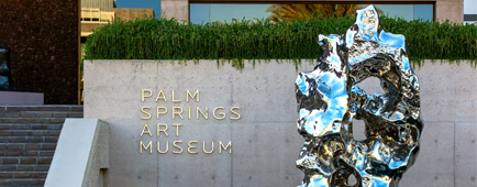 A metal sculpture sits in front of a Palm Springs Art Museum sign. A metal sculpture sits in front of a Palm Springs Art Museum sign.