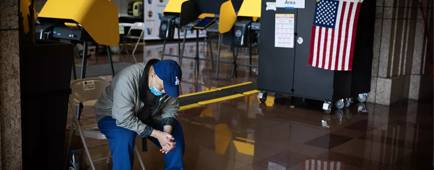 A man in a blue cap looks down while seated in a room with voting stations with yellow privacy screens
