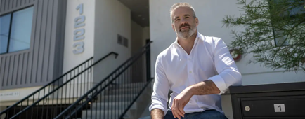 A man in a white shirt sits in front of an apartment building 
