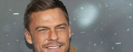 Alan Ritchson smiling in a tan coat  with a flipped-up collar