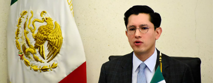 A young man with dark hair and glasses, in dark suit and light blue tie, seated near green-white-and-red flags