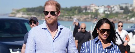 Prince Harry and Meghan Markle wear sunglasses and blue button down shirts as they walk hand in hand.