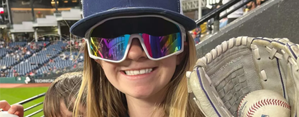 A girl in a Guardians hat and sunglasses smiles while holding a baseball in a glove. A boy peeks around her with another ball