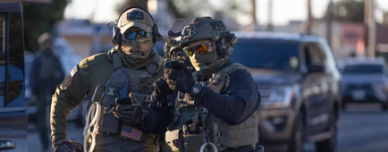 Armed federal agents with masks, hats and sunglasses looking at their phone screens while standing in a street