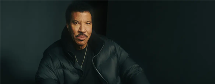 Lionel Richie in a dark outfit poses with his hands on his lap against a dark backdrop. 